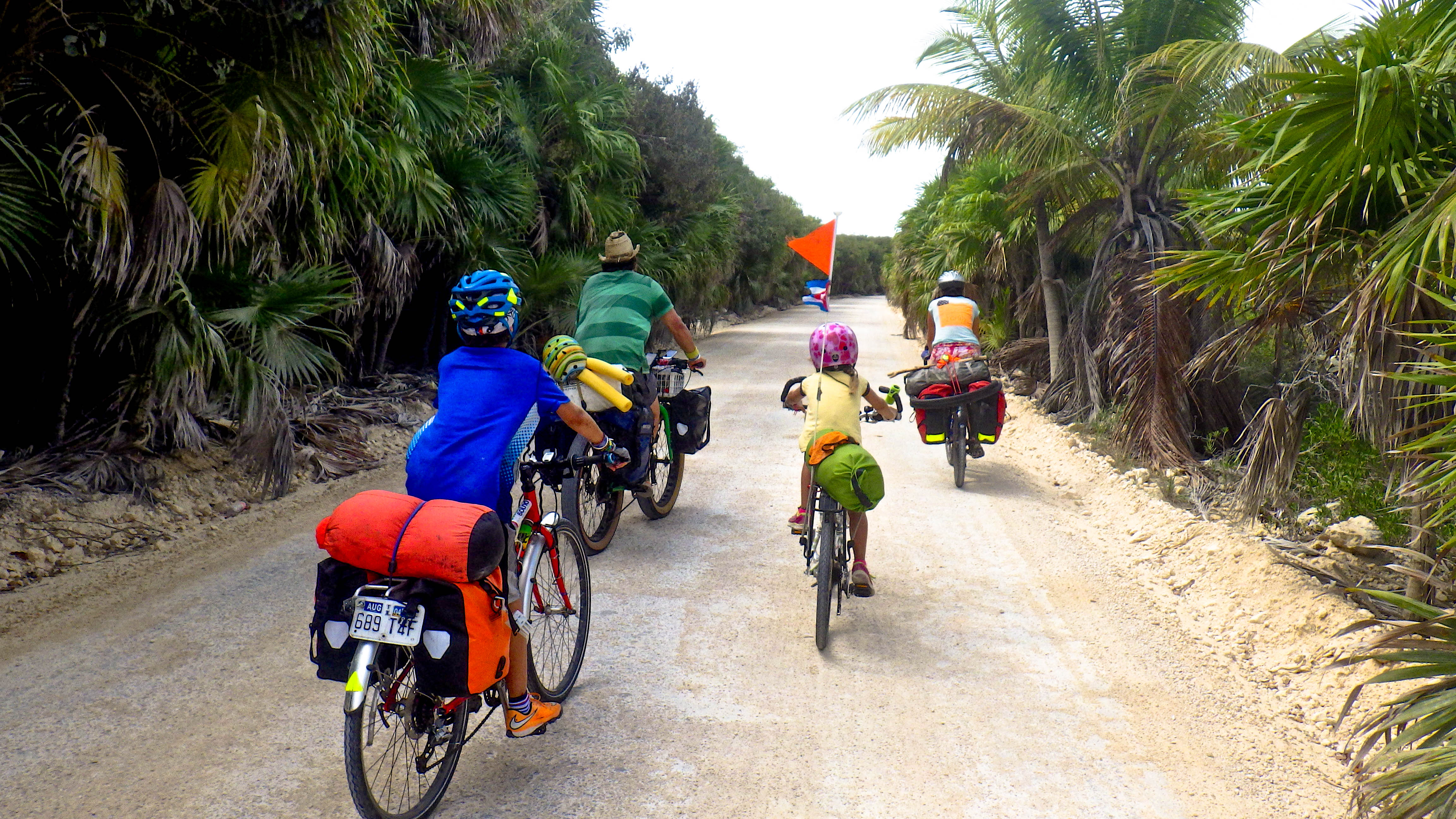 A traveling family cycles on a dirt road surrounded by tropical plants, showing parents who share their wanderlust with their kids. (image © Eva Boynton)
