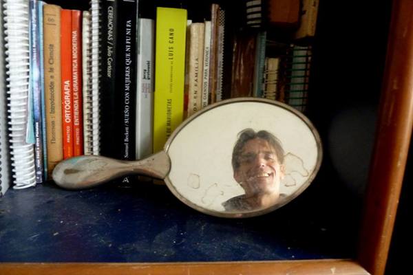 Selfie as a still life full of creative expression shows a man's photo in a hand mirror on a shelf of books. (Image © Javier Maubecin)