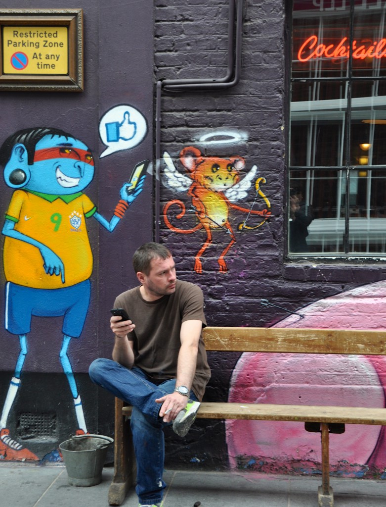 Man using smart phone on bench next to wall with creative street art portraits. (Image © Sheron Long)