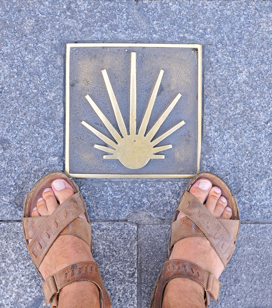 Feet in sandals standing over a sign of the Camino de Santiago, showing the different routes that cause travel inspiration. (image