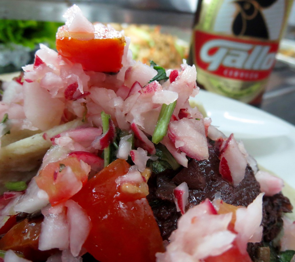 Close up of a moronga (blood sausage) taco with tomatoes and onions, and a Gallo beer bottle in the background., a tasty memory recalled during the post-vacation blues. (Image © Sally Baho)