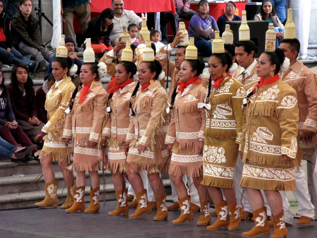 Women and men dressed in cowboy-like outfits balancing bottles on their heads, showing influences of border culture on traditional Mexican dances. (Image  © Eva Boynton)