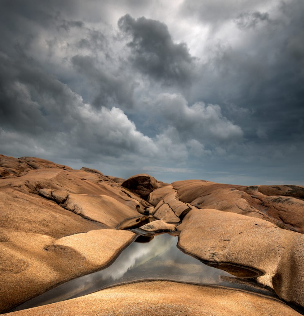 Sand colored stones, water, and storm clouds in Sweden, landscape photography that provides a virtual journey and a celebration of Earth Day. (Image © Claes Thorberntsson.)