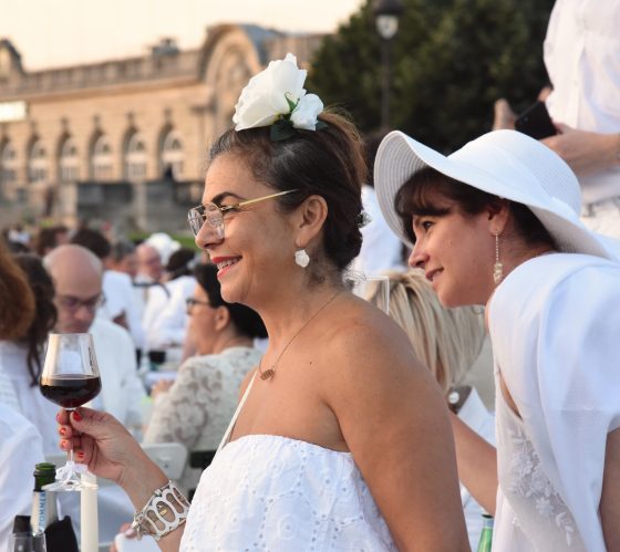 Two women in white with a glass of red wine at the Paris Dîner en Blanc, enjoying the cultural traditions of the Dinner in White. (Image © Meredith Mullins.)