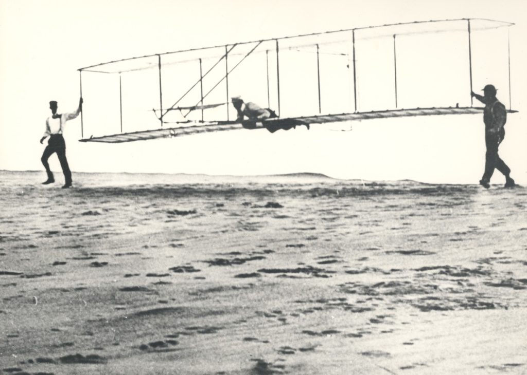 Men with an early flying machine evoke aviation innovations and flights of fancy. [Image public domain]