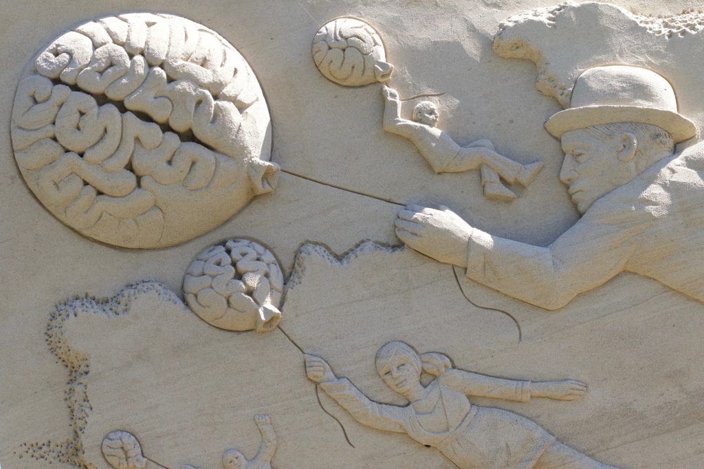 A sand sculpture of people borne aloft by balloons that resemble brains symbolizes the brain’s power to use hindsight to boost our ability to learn a language. (Public domain image by FotoEmotions/Pixabay)