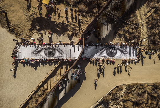 Artivist JR organizes a picnic on both sides of the border fence near Tecate Mexico, showing cultural diversity and social awareness and answering the question can art change the world. (Image © JR.)