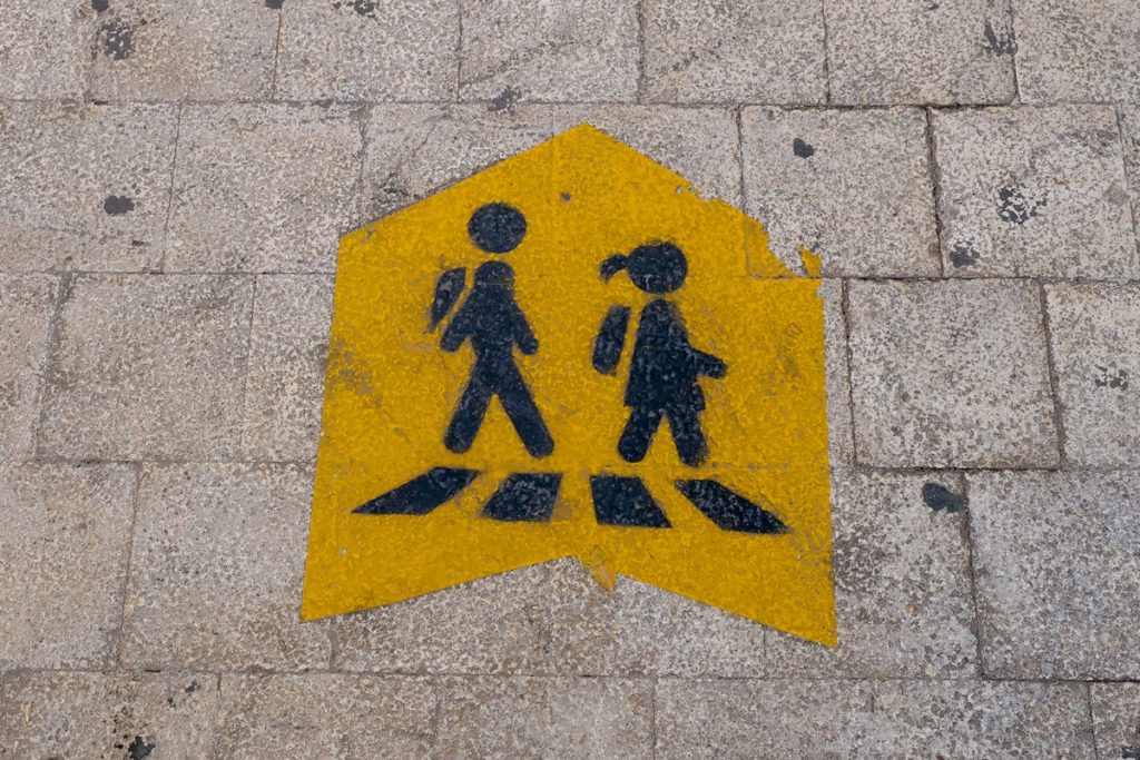 This sidewalk sign shows a female and male student in profile and with backpacks as it points its way to a nearby school and is part of a series of school zone signs from different cultures. Image © jansmartino/iStock .