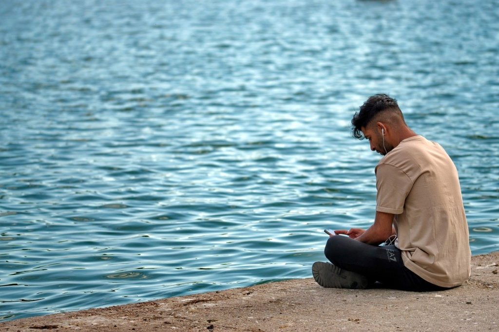 A man sits by the water, listening but also wearing earphones, a reminder of why we need World Listening Day to reunite our global community in the act of mindful listening. (Image by Pixabay)